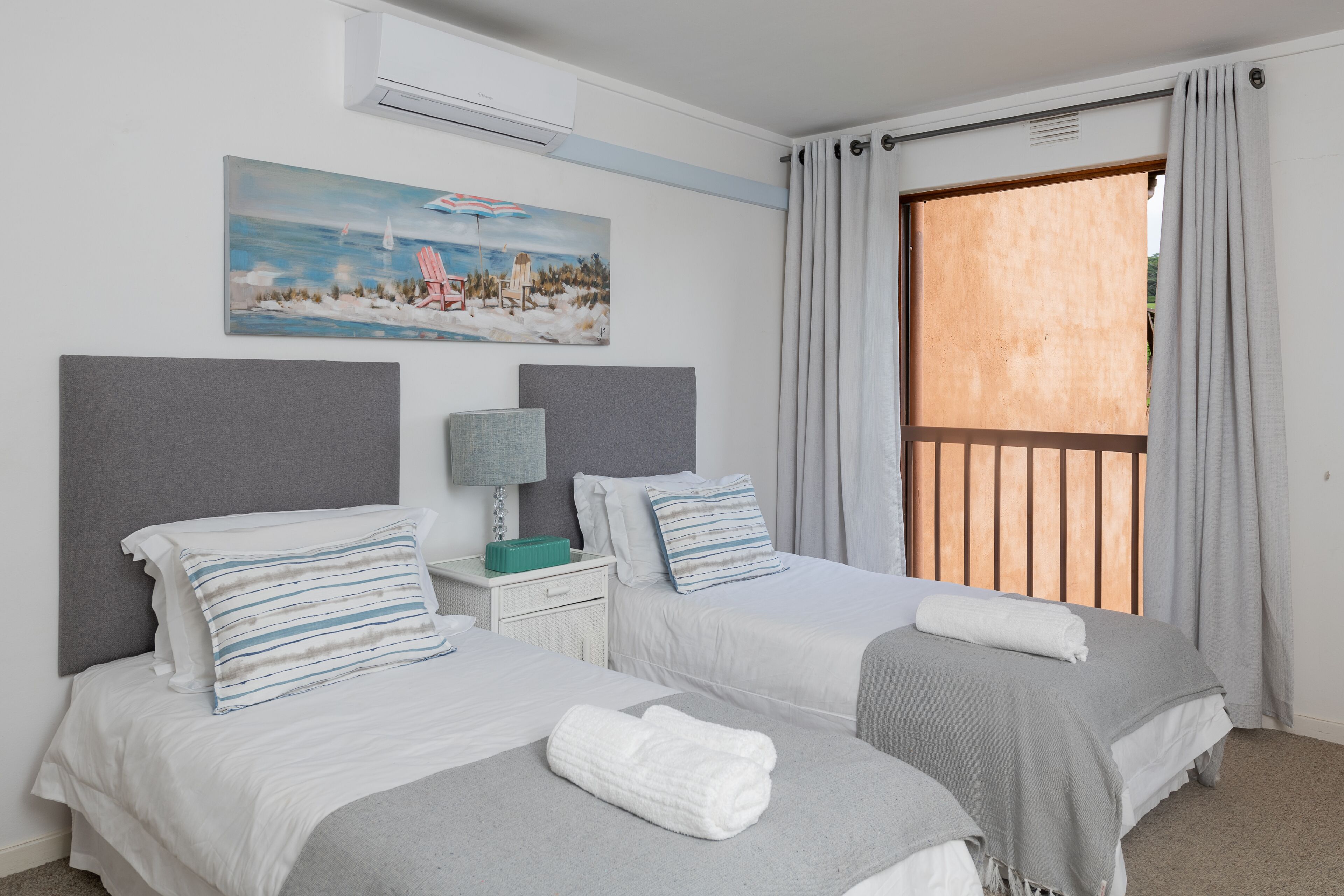 A hotel room with two beds, a painting of a beach scene, a window with curtains, and a nightstand with a lamp.