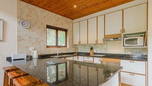 Cozinha com ilha central, banquetas de madeira, armários brancos, micro-ondas e janela com persianas.