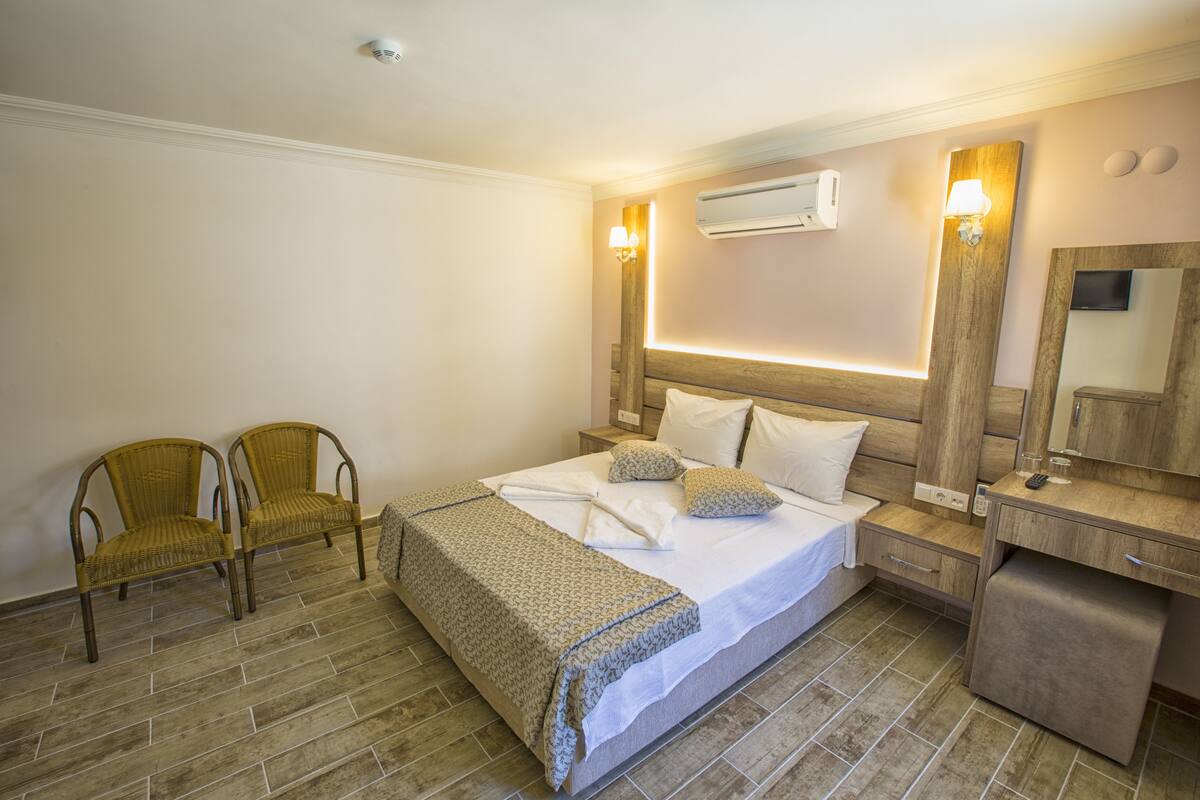 A hotel room with a bed, two chairs, a bedside table, a mirror, and an air conditioning unit.