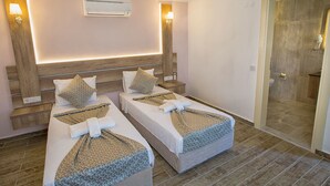 A hotel room with two beds, each with a towel and a blanket, a wooden headboard, a wall-mounted air conditioner, and a shower area visible through a door. - Kas Otel (Kas)