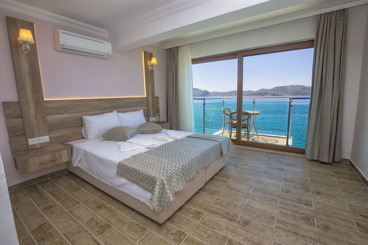 A bedroom with a large bed, a nightstand, a wall-mounted air conditioner, a window with a view of the sea, and a balcony with a table and chairs.