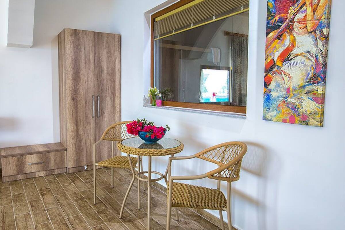 A room with a wooden wardrobe, a small table with a vase of flowers, wicker chairs, and a large window with a view.