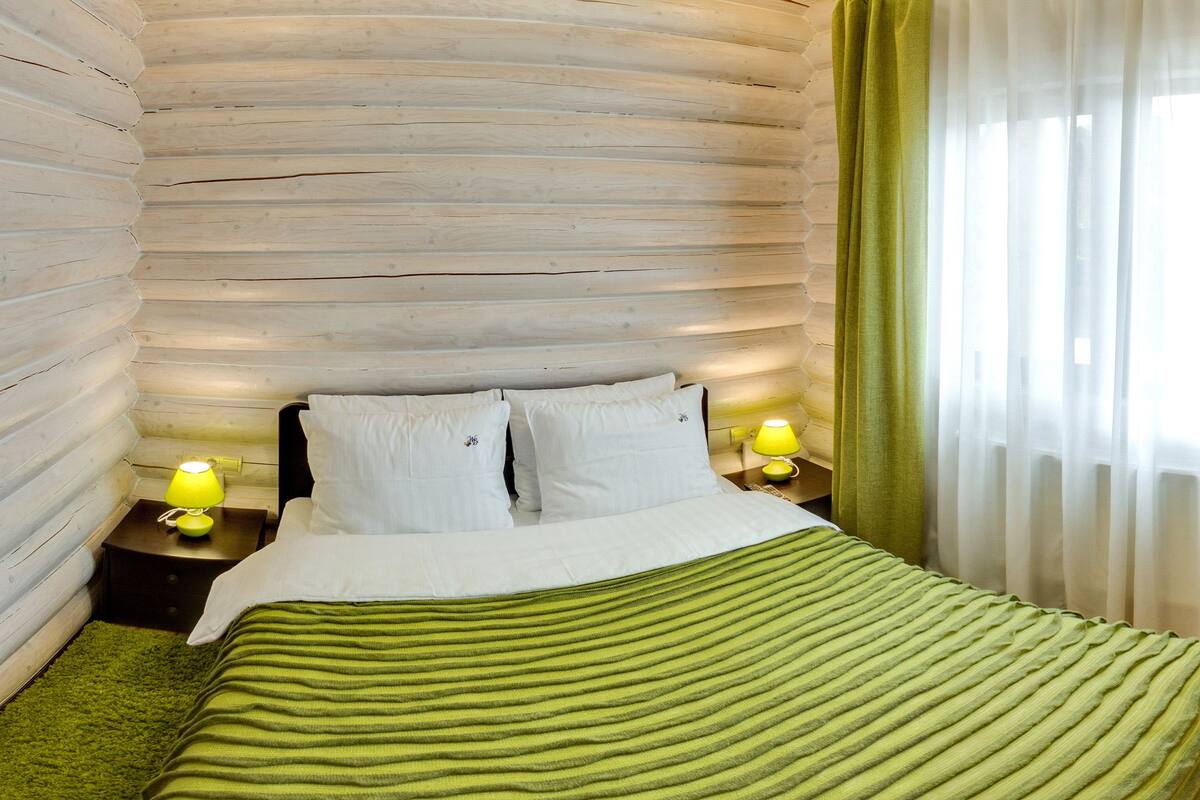 Habitación con pared de troncos, cama con ropa blanca, dos lámparas de noche y una manta verde a rayas.