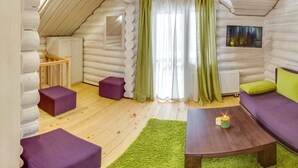 Ein gemütliches Blockhaus-Interieur mit einer Couch, zwei violetten Hocker, einem Holztisch mit Kerzen und einem Fernseher an der Wand.