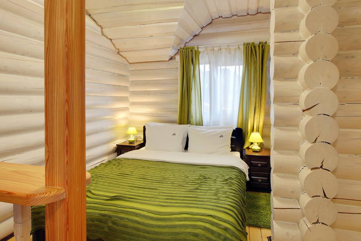 Una habitación con techo de listones de madera, una cama con colcha verde a rayas, dos almohadas blancas y una mesita de noche con lámpara.