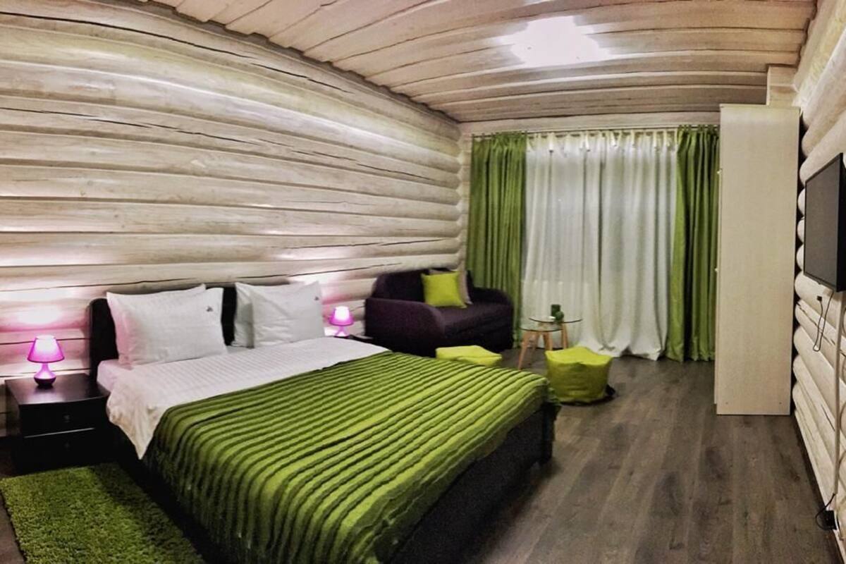 Un dormitorio con pared de troncos de madera, una cama con ropa de cama verde, una mesita de noche con lámpara, una silla, un televisor montado en la pared y una ventana con cortinas.