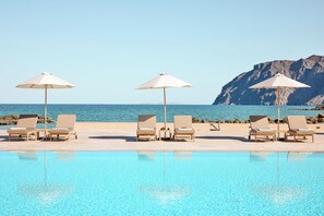 A poolside area with lounge chairs and umbrellas, a clear view of the sea, and a rocky cliff in the background.