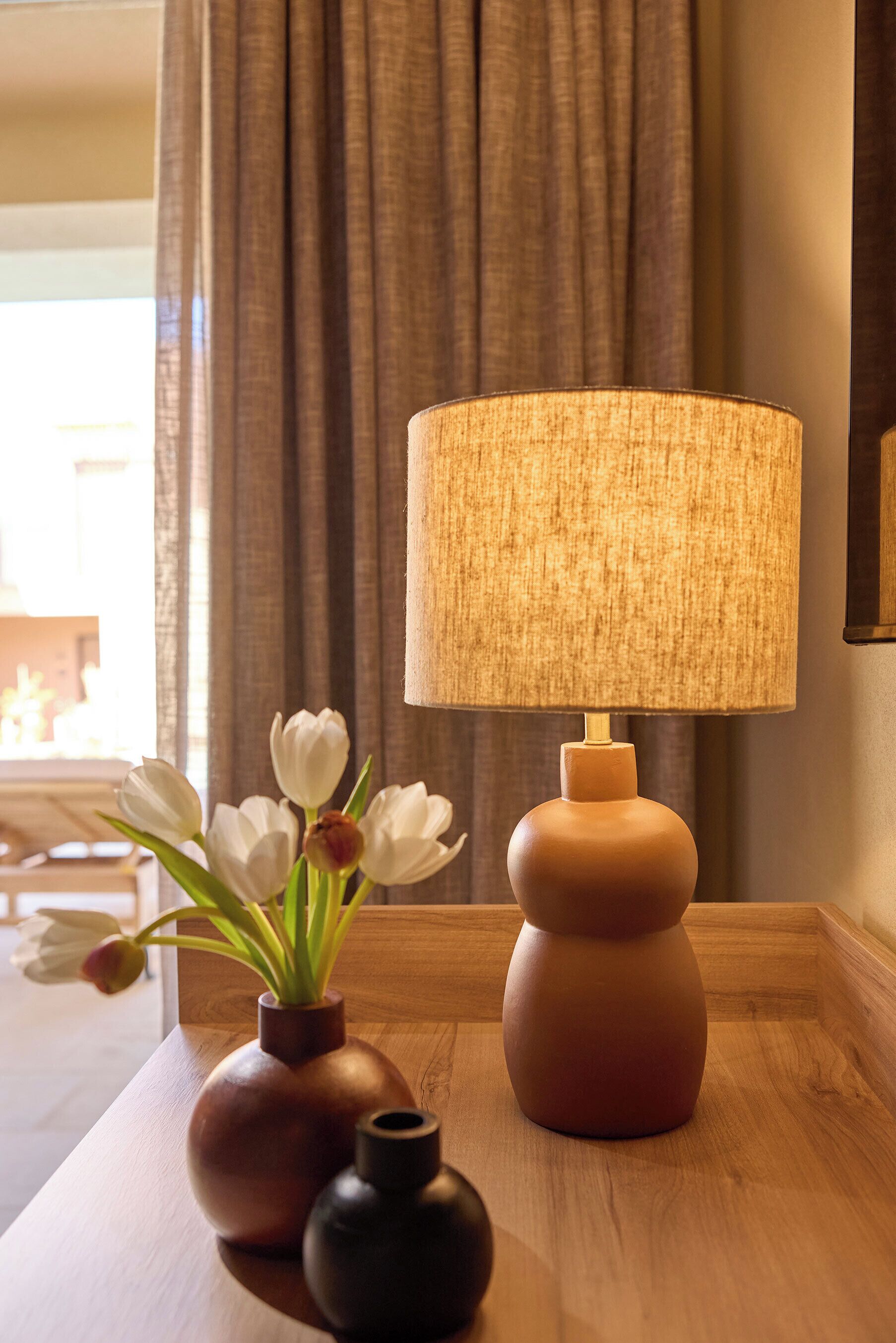 Une table de chevet en bois, une lampe, un vase avec des fleurs blanches et un vase plus petit.