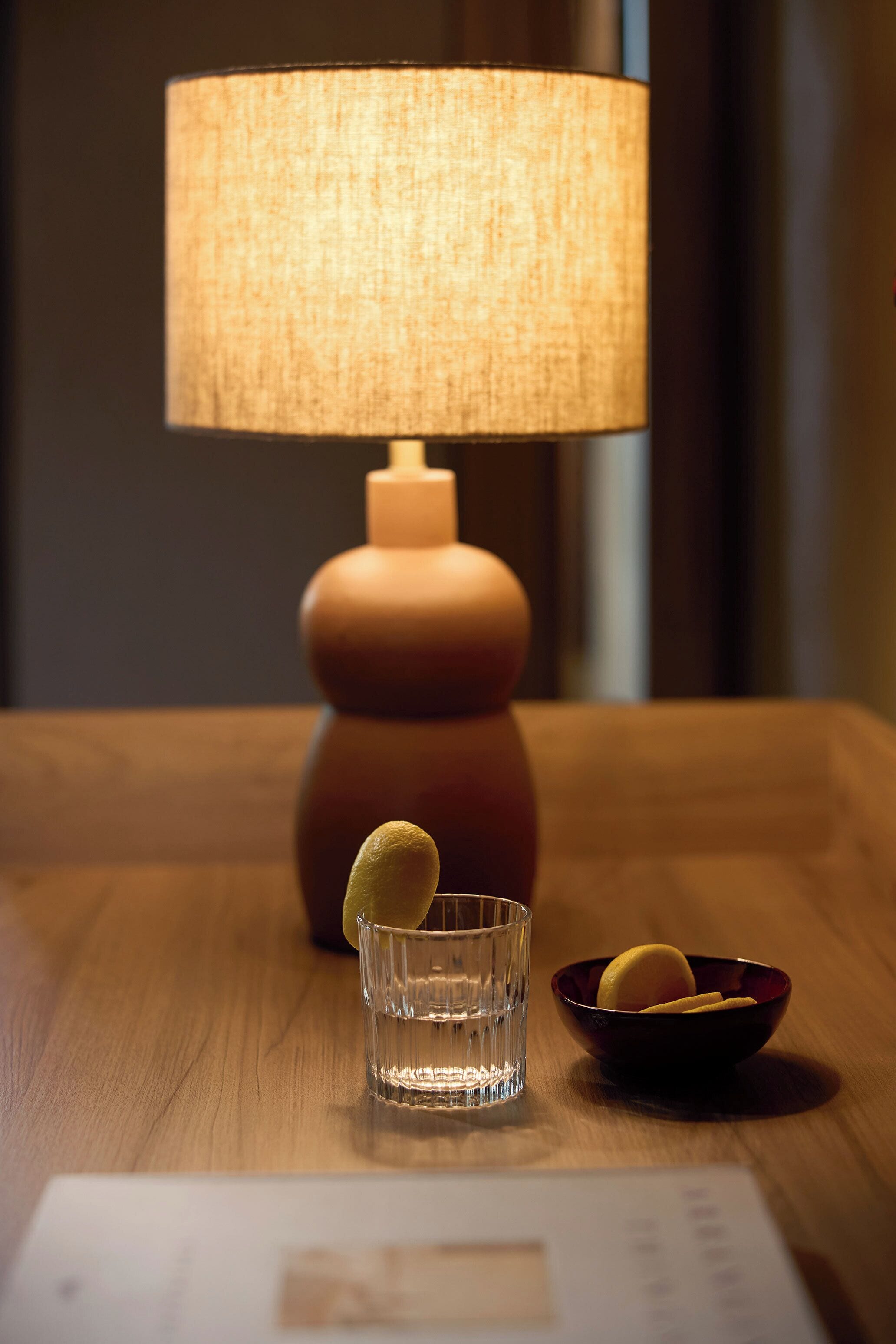 A wooden table with a lamp, a glass, and a bowl containing a lemon slice.
