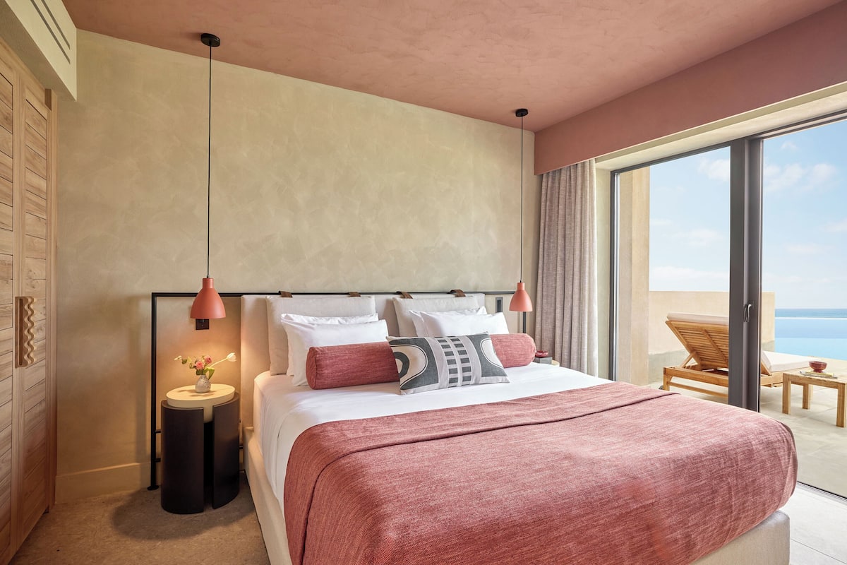 Ett modernt hotellrum med en stor säng, ett nattduksbord med en lampa och utsikt över havet genom en skjutdörr av glas.