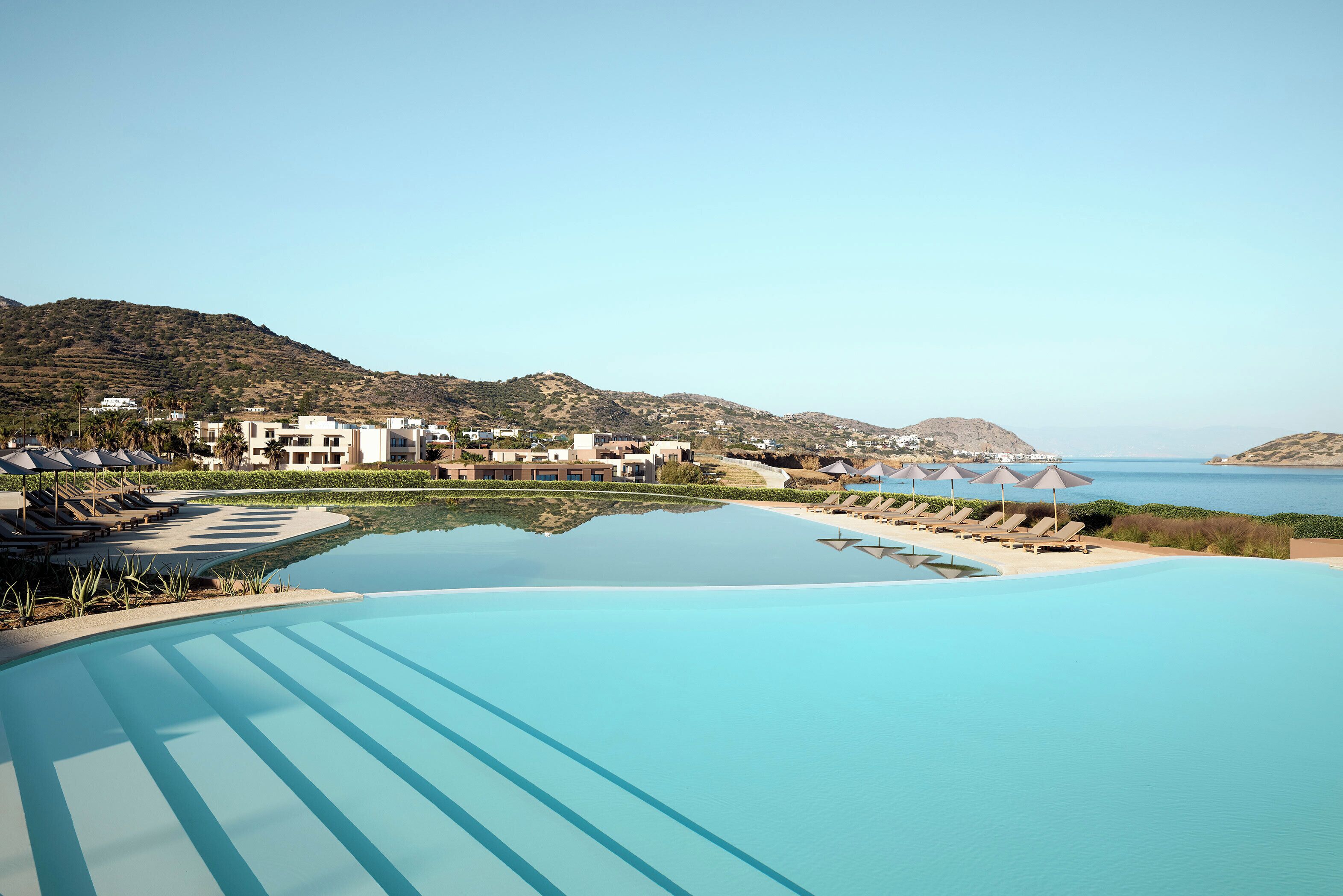 Un resort con una amplia piscina infinita, tumbonas y vistas al mar y las colinas.