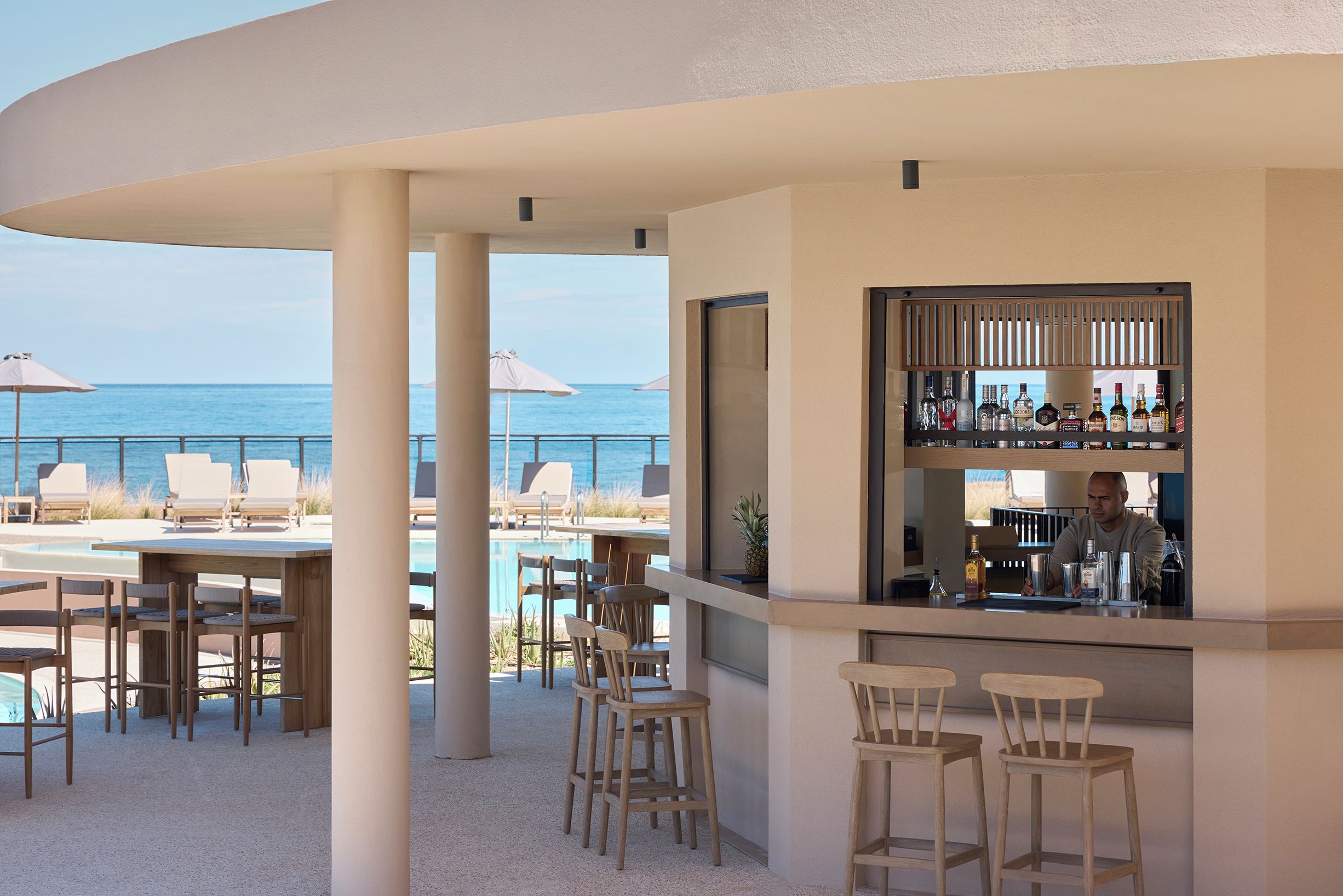 A poolside bar area with a view of the ocean, bar stools, and lounge chairs.