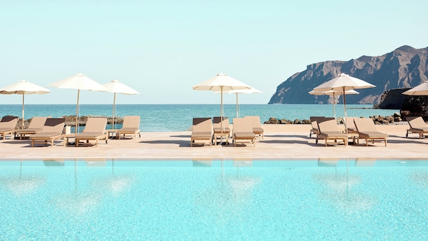 A pool area with lounge chairs and umbrellas, a clear sky, and a mountainous backdrop.