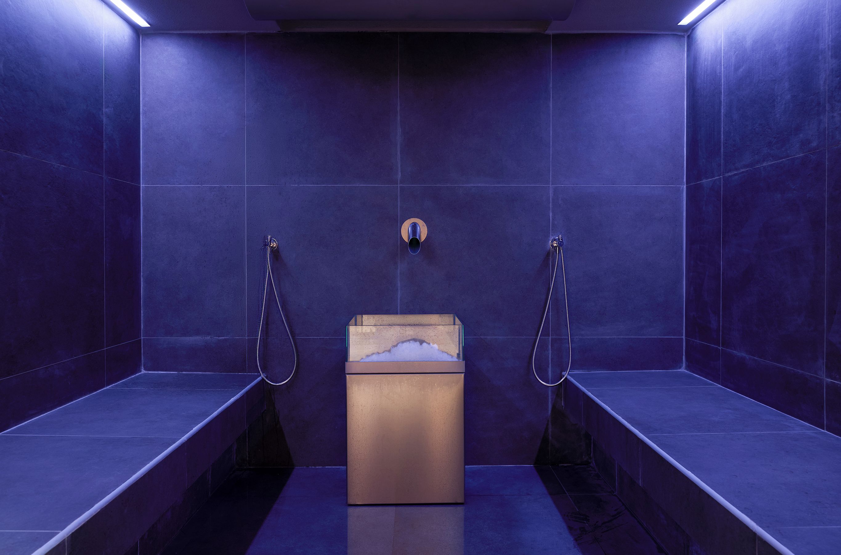 A modern bathroom with dark blue tiles, a central sink with a soap dispenser, and two showerheads.