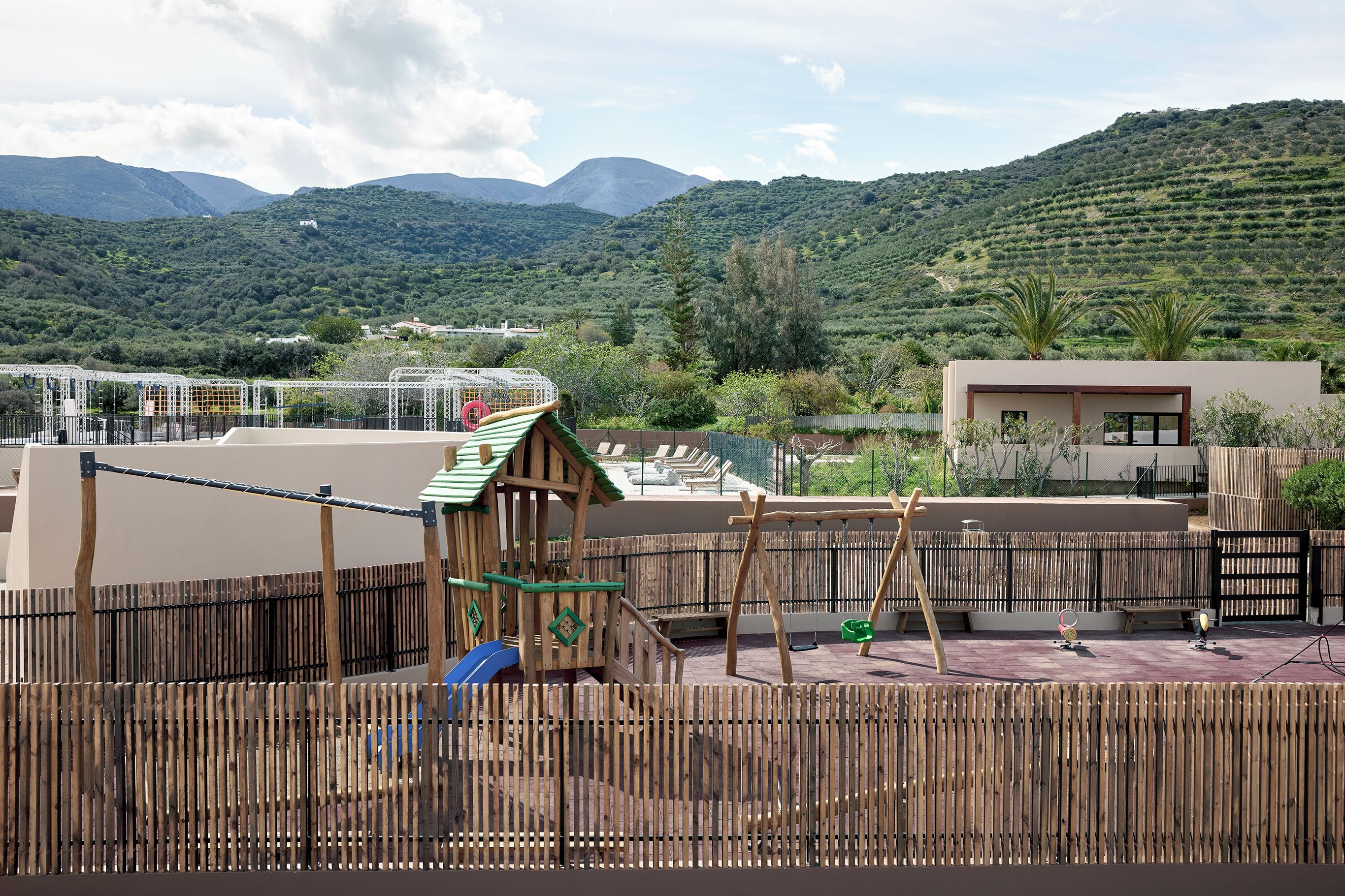 A playground with a wooden play structure and a swing set, surrounded by a bamboo fence.