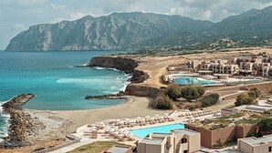 A coastal resort with a beach, swimming pools, and a mountainous backdrop.