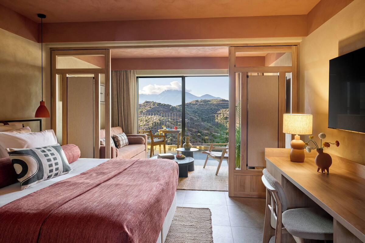 Ett hotellrum med en stor säng, ett skrivbord, en stol, en TV och en balkong med utsikt över bergen.