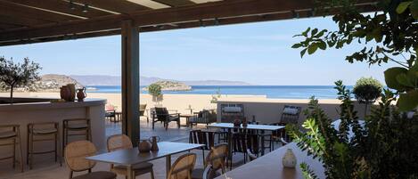An outdoor patio with tables and chairs, overlooking a beach and the sea.