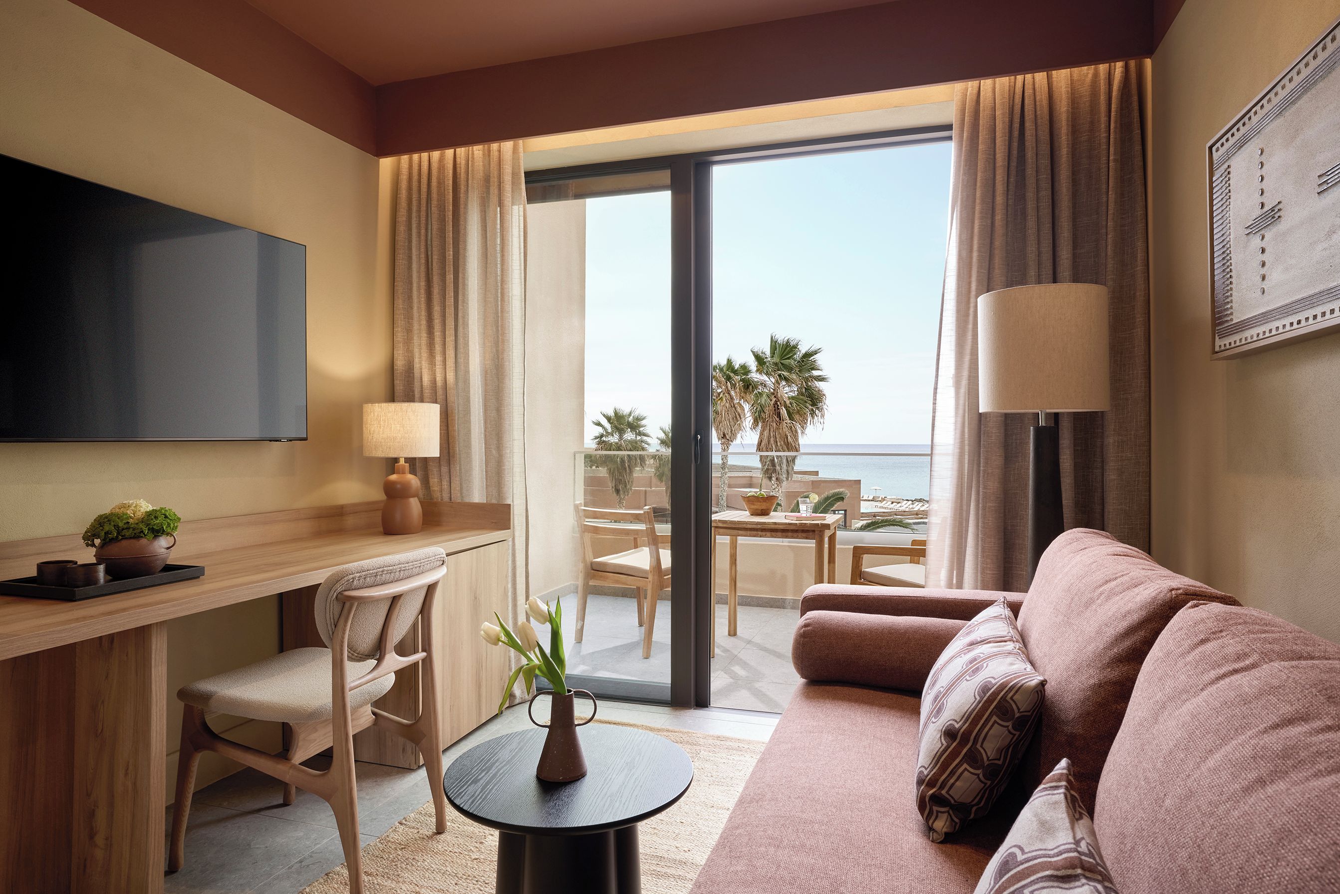 Une chambre d’hôtel équipée d’un téléviseur à écran plat, d’un canapé, d’une table à manger avec des chaises, d’une petite table ronde agrémentée d’un vase, et offrant une vue sur l’océan depuis les portes du balcon.