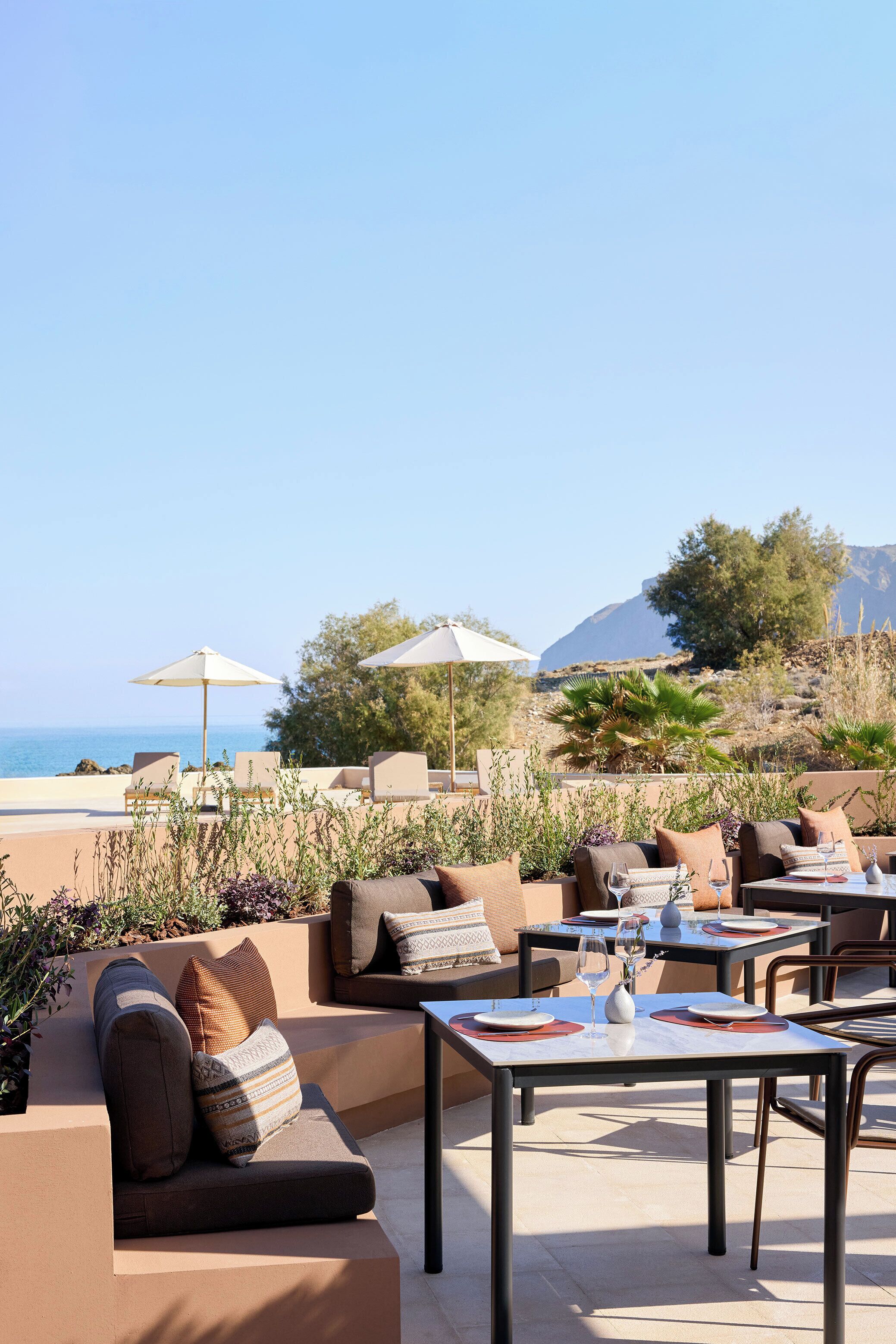 An outdoor seating area with tables and chairs, some with umbrellas, overlooking a body of water and hills.