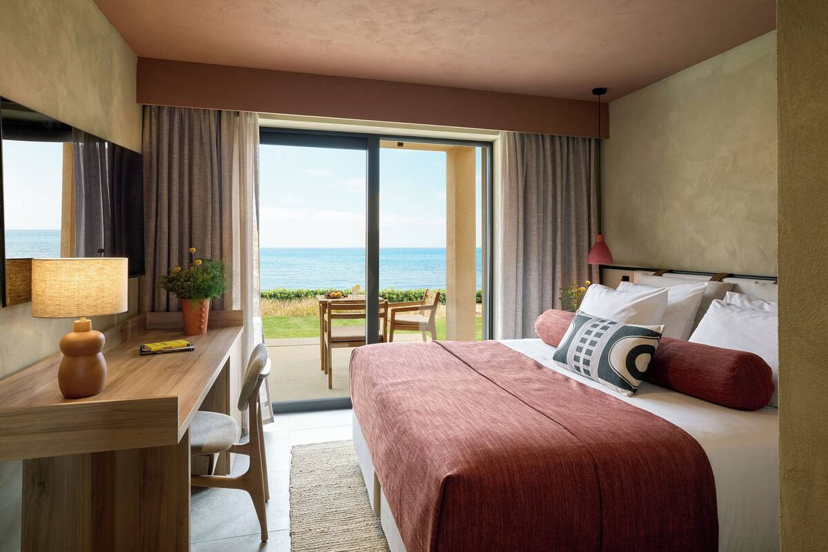 Ett hotellrum med en säng, ett skrivbord, en stol, en lampa och utsikt över havet genom en skjutdörr.