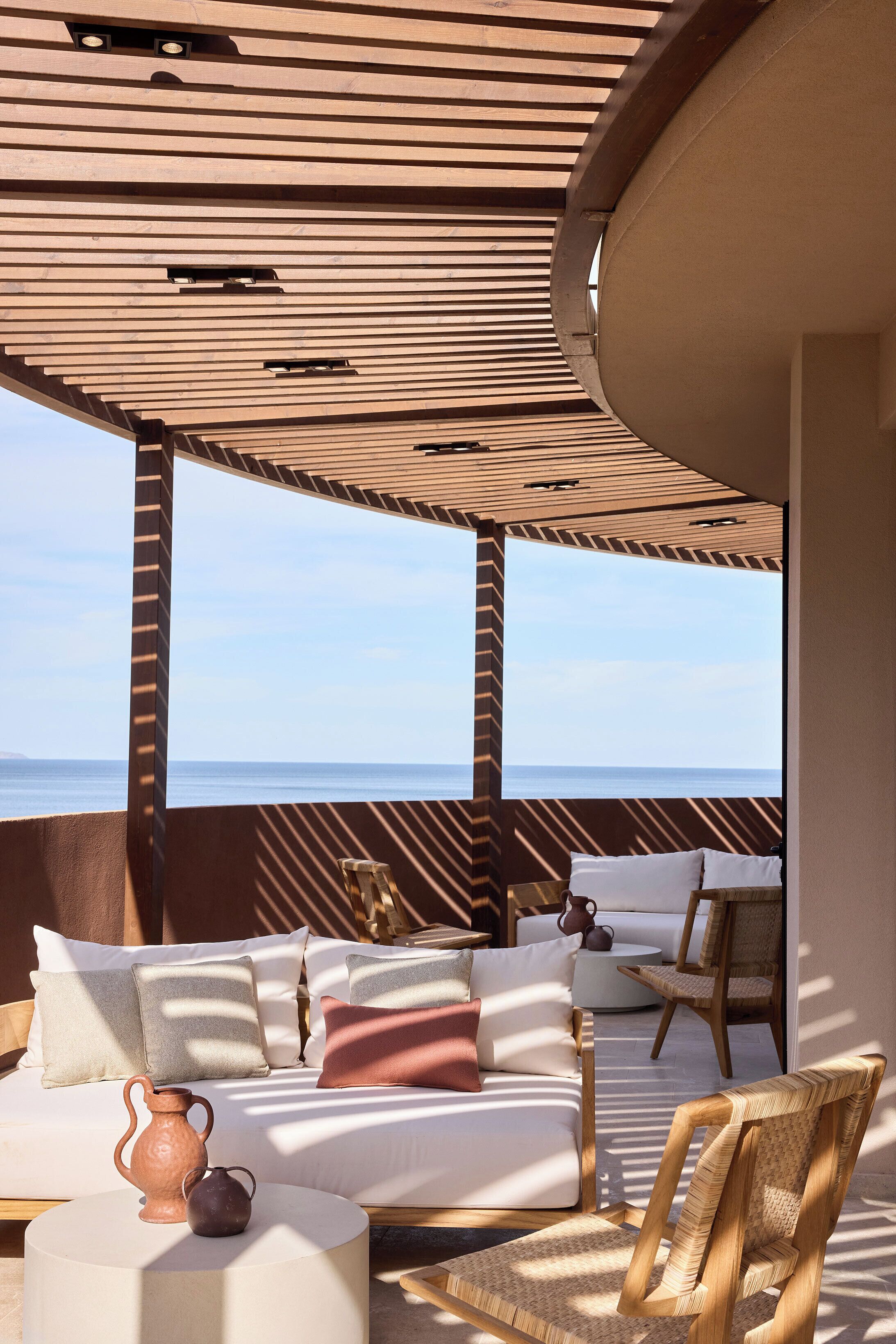 A sunlit outdoor seating area with a view of the sea, featuring a sofa, chairs, and a small table with pottery.