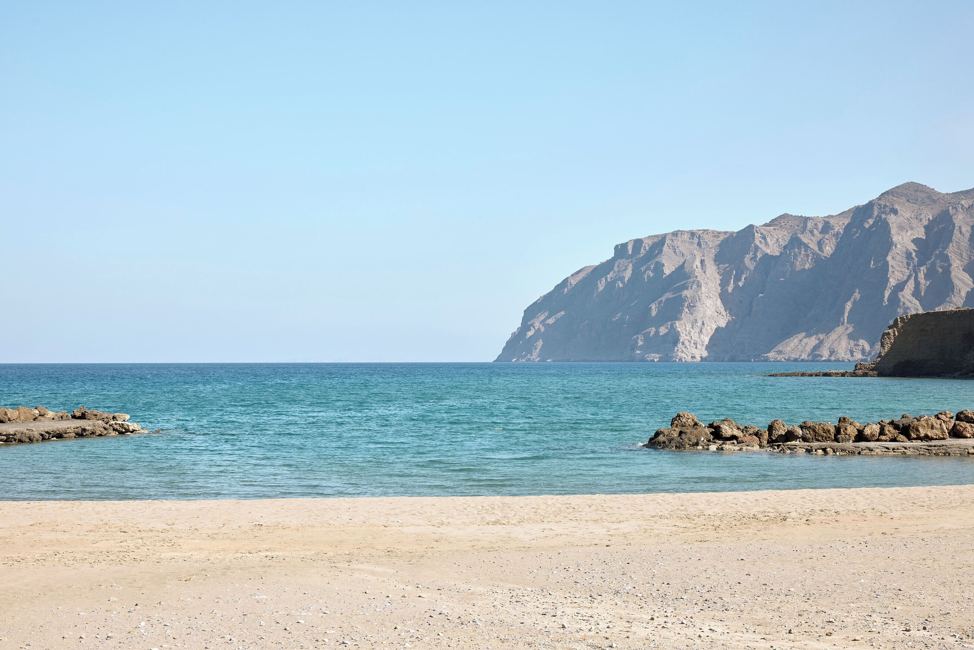 Une plage à l’eau d’un bleu cristallin, bordée d’affleurements rocheux et dominée par un paysage montagneux.