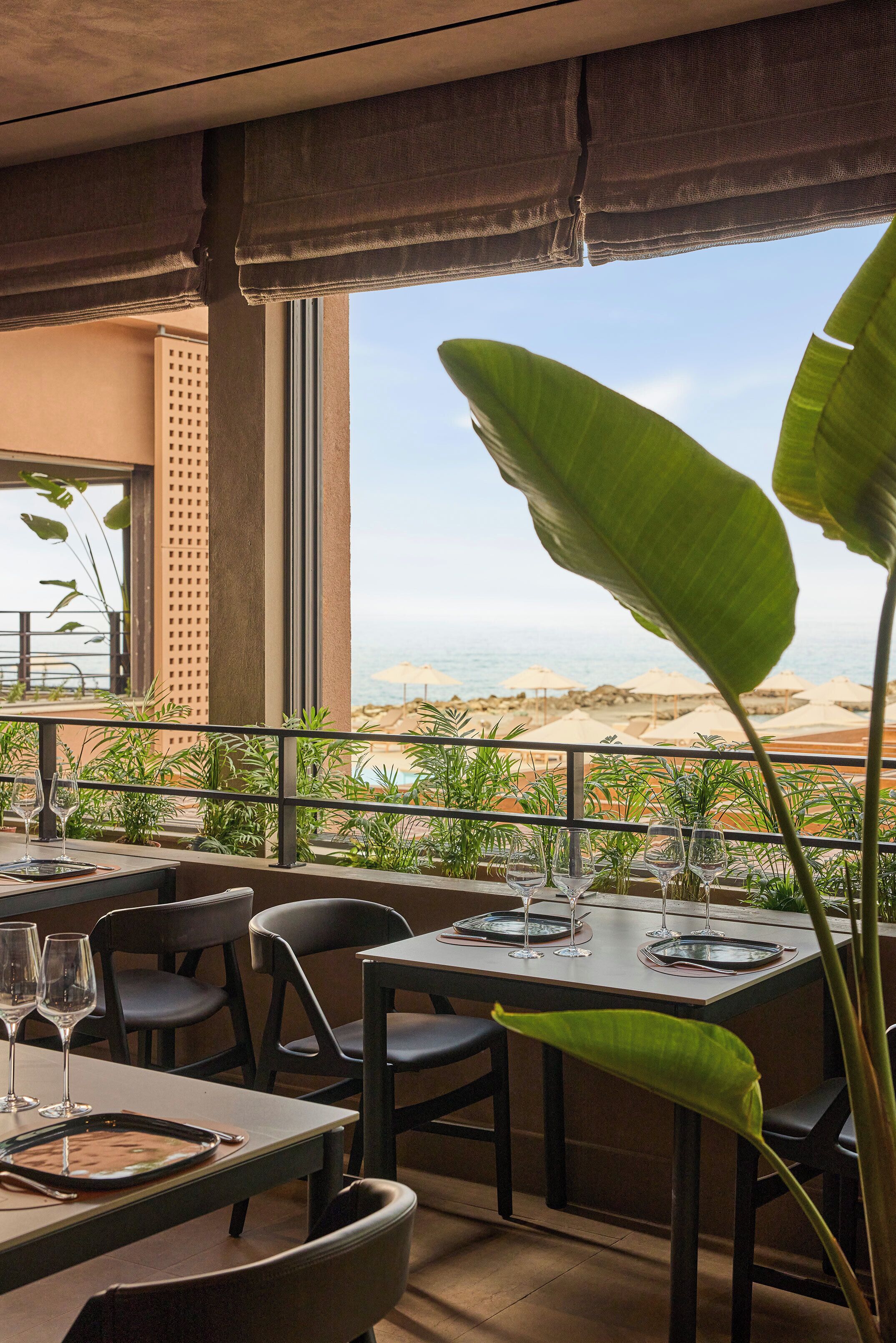Un restaurante con mesas dispuestas para comer, grandes plantas verdes y vistas a la playa y al océano.