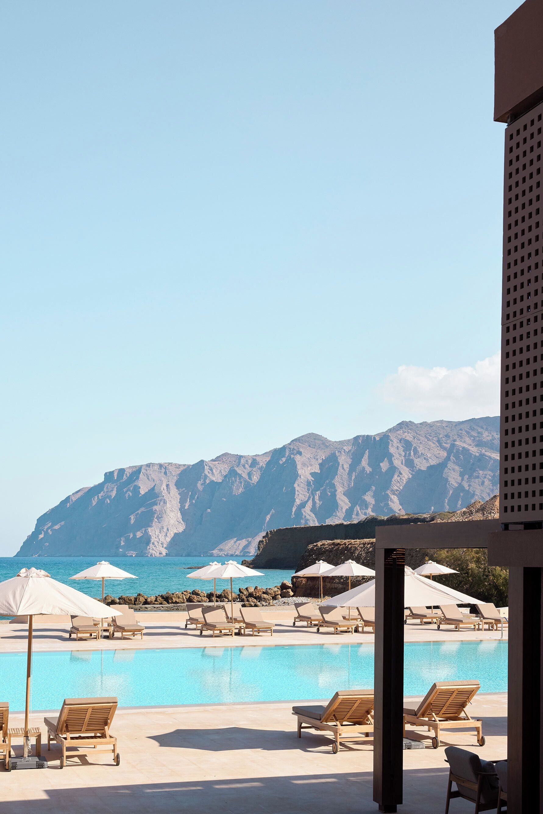 Zona junto a la piscina con sillas de descanso y sombrillas, con vistas a un paisaje montañoso y al mar.