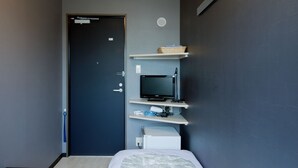 Une chambre d’hôtel de taille réduite, comprenant un lit, une petite table, un téléviseur posé sur une étagère et une porte.