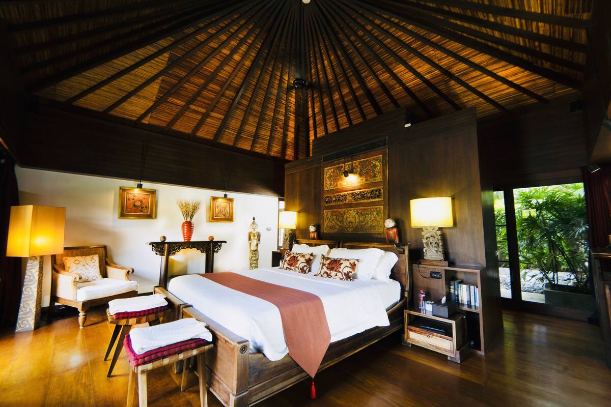 A spacious bedroom with a large bed, wooden furniture, and a thatched ceiling.