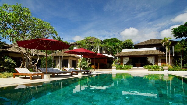 A pool area with lounge chairs and red umbrellas, surrounded by tropical vegetation and buildings. - Tamarind Exclusive Villa (Sattahip)