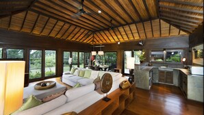 A spacious living area with a wooden ceiling, large windows, a sofa with cushions, a coffee table, and a kitchen area with a sink and countertop. - Tamarind Exclusive Villa (Sattahip)