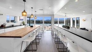 A modern kitchen with a long island, white bar stools, pendant lighting, and large windows offering a view of a lake.