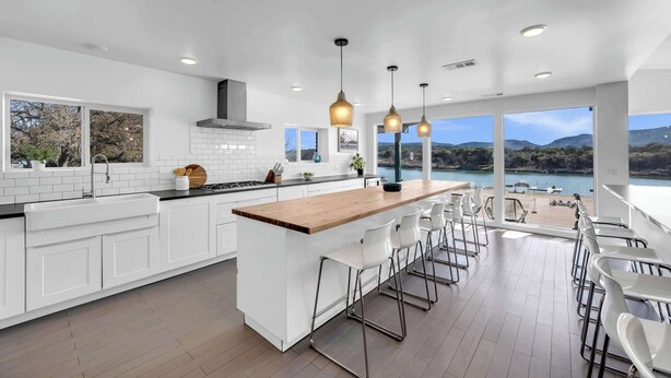 A modern kitchen with white cabinetry, a wooden island, and bar stools. It features large windows with a view of a lake and mountains.