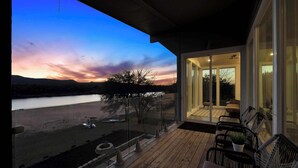 A wooden deck with outdoor furniture, a glass door leading to an interior space, and a view of a lake at dusk.