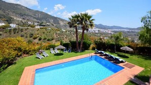 A swimming pool with lounge chairs and a palm tree, surrounded by a well-maintained lawn and a view of a hilly landscape. - Great Villa ideal for groups: swimming pool, wifi, air conditioning, sea view. (Frigiliana)