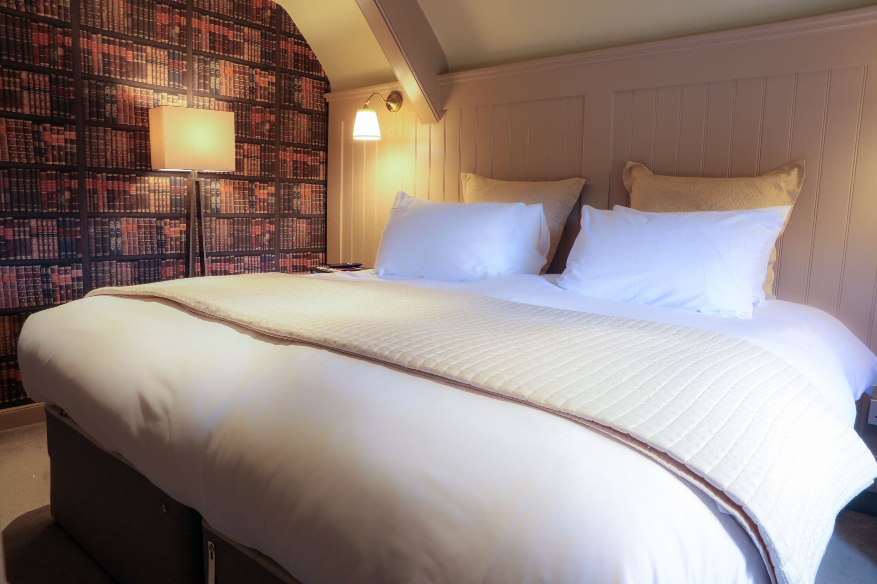 A large bed with white linens, a bedside lamp, and a bookshelf in the background.