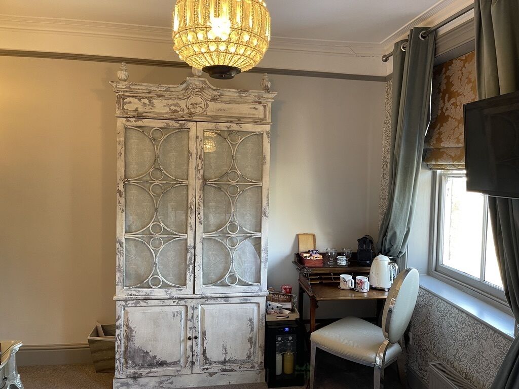 A room with a vintage cabinet, a table with a kettle, and a chair by a window.