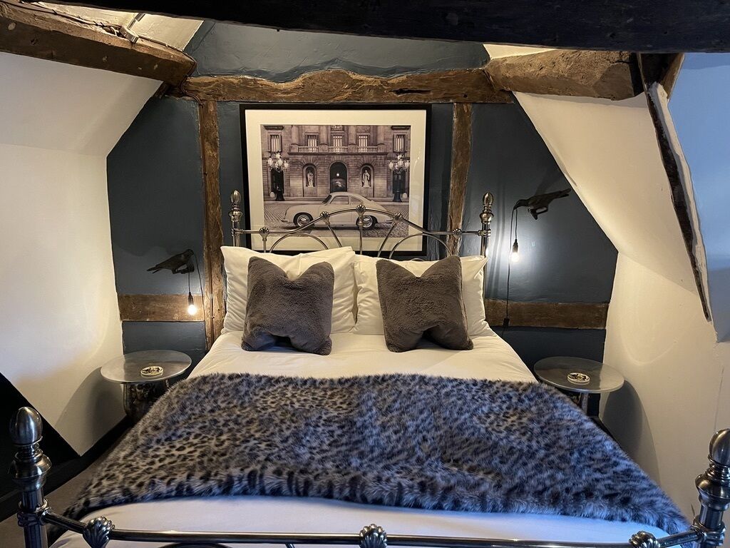 Una cama con cabecera, dos mesitas de noche, un cuadro enmarcado en la pared y una ventana con cortinas.