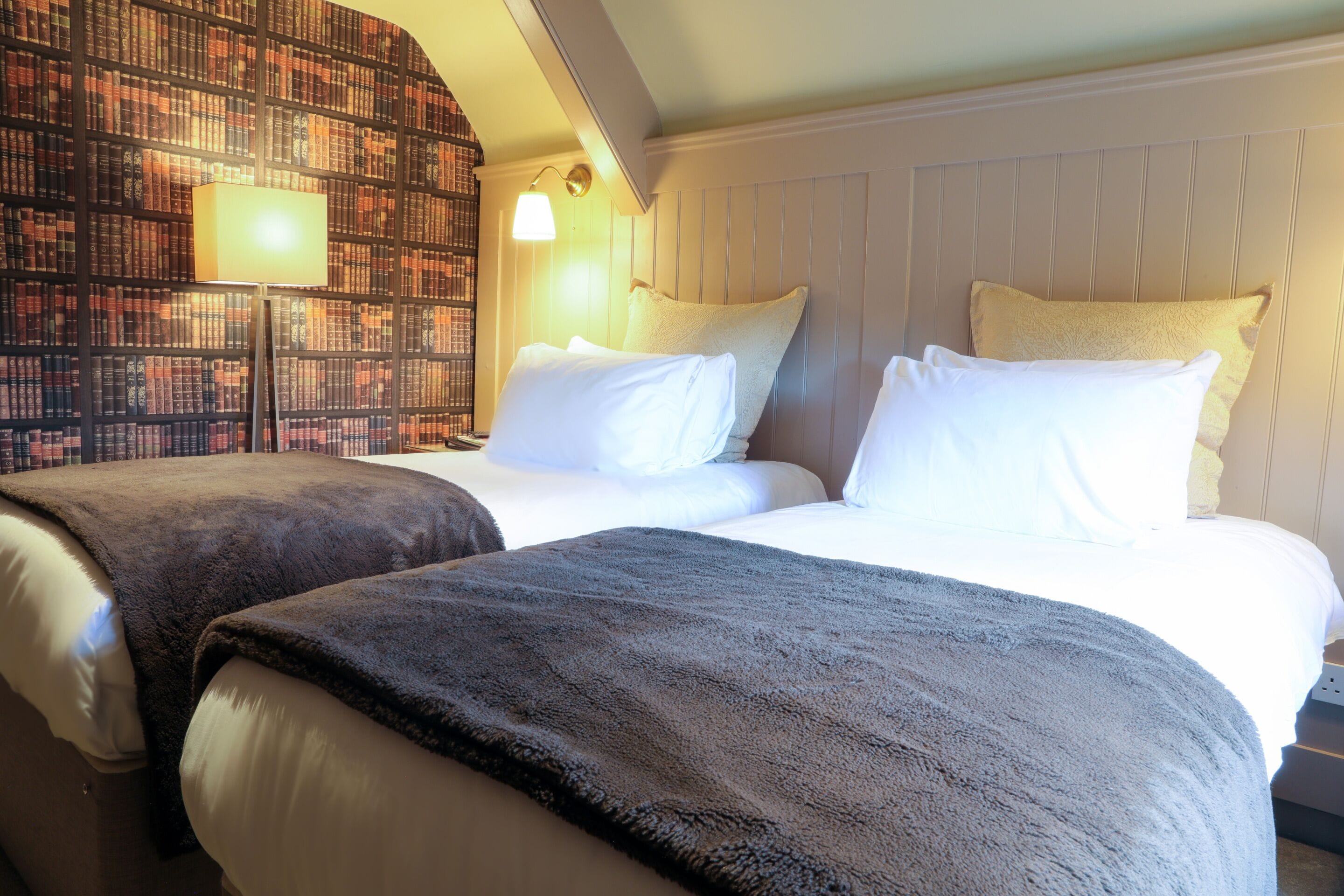 A hotel room with two beds, a bookshelf wall, and a lamp.