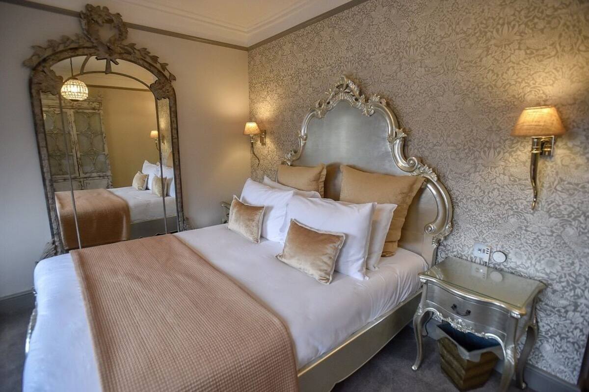 A hotel room with a large bed, ornate headboard, a mirror, a nightstand, and a wall with patterned wallpaper.