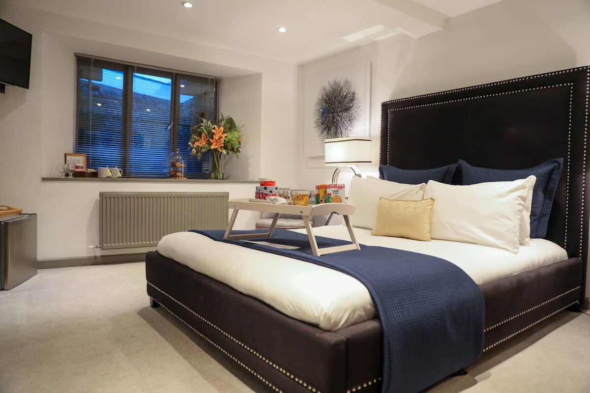 A modern bedroom with a large bed, a bedside table, a window with blinds, a radiator, and a wall-mounted TV.