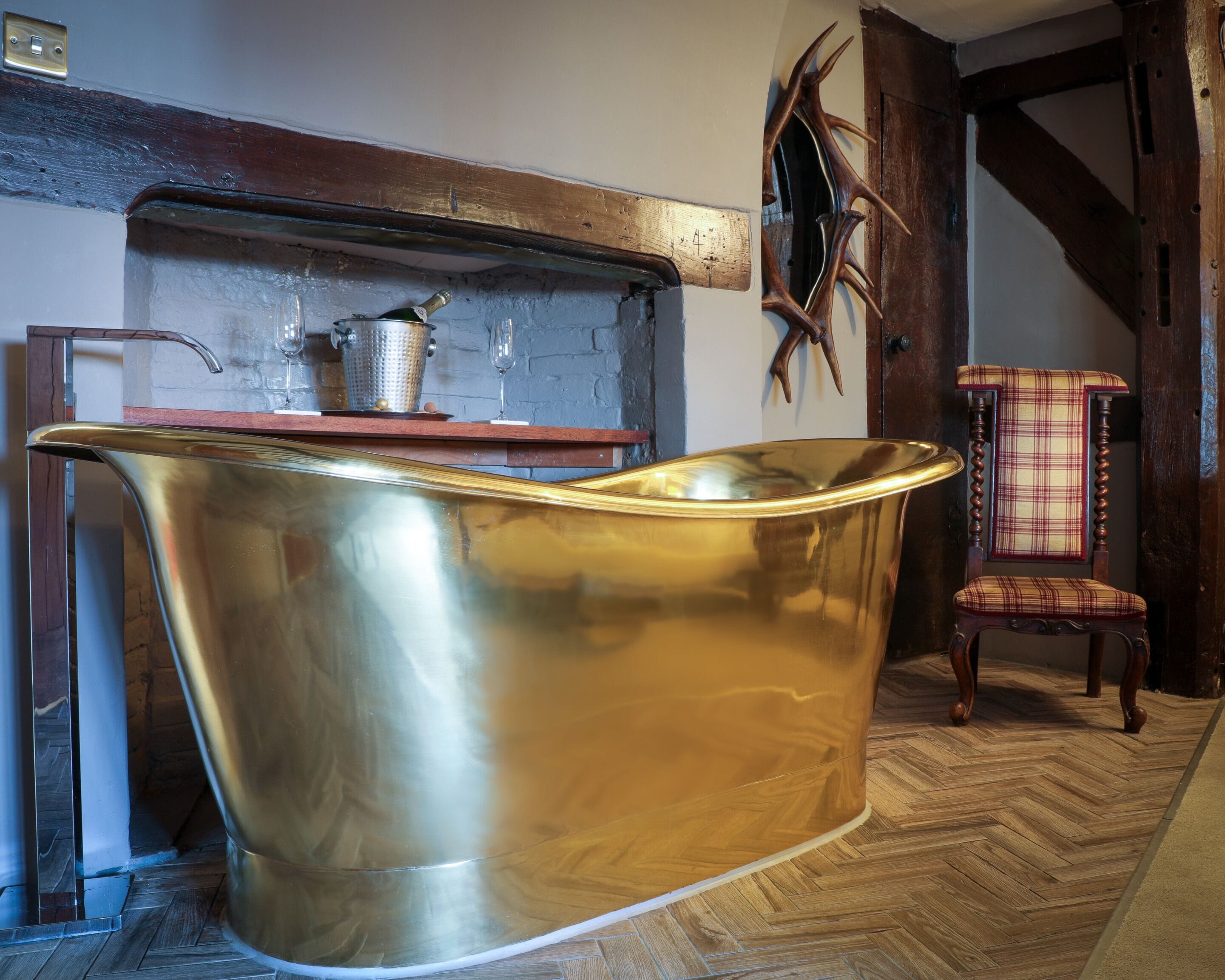 Una habitación con una bañera metálica grande y brillante, una silla de madera con cojín escocés y una puerta de madera rústica.