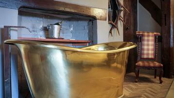 Una habitación con una bañera metálica grande y brillante, una silla de madera con cojín escocés y una puerta de madera rústica.