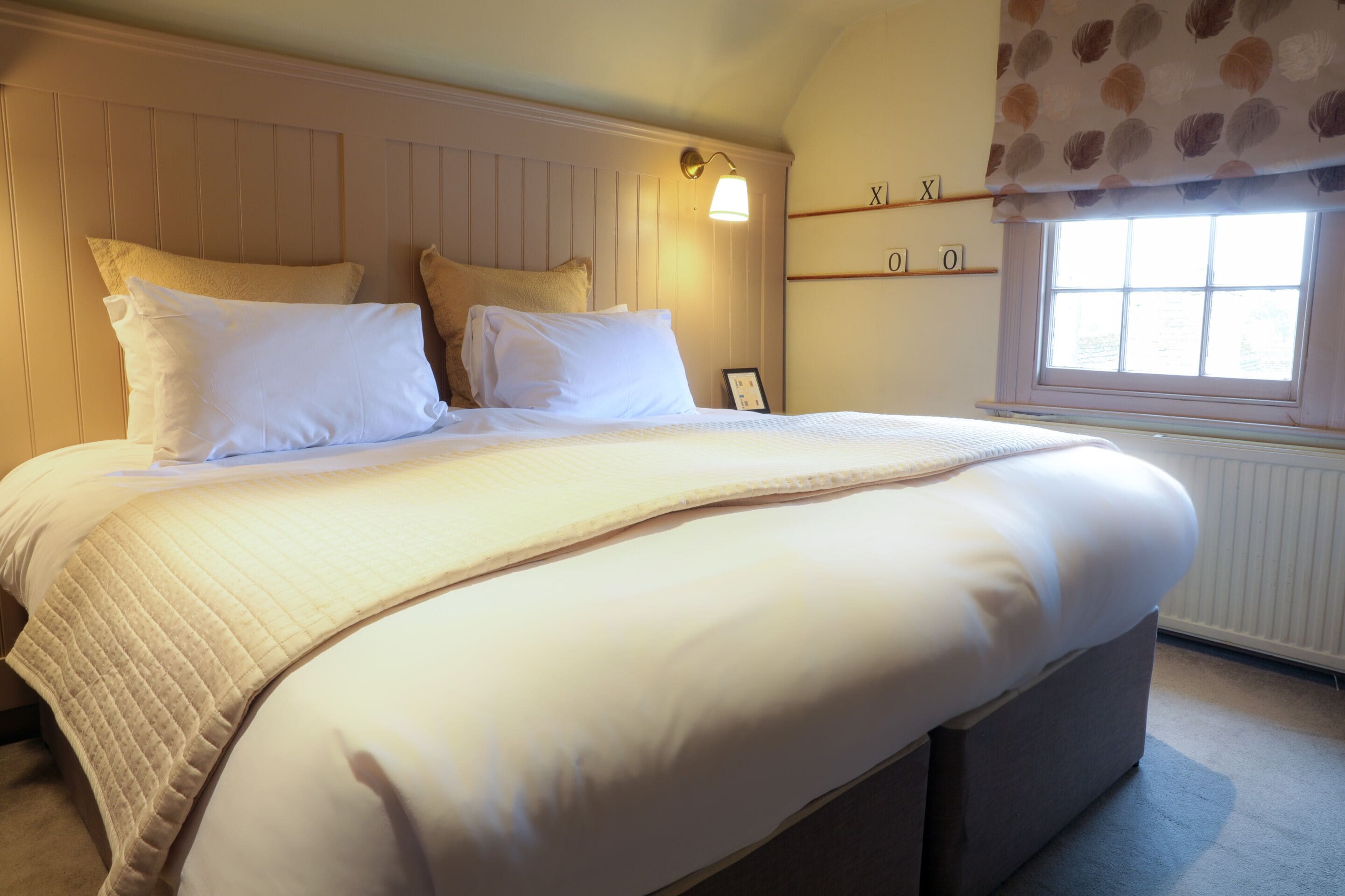 A neatly made bed with a quilted cover, a bedside table with a lamp, and a window with a curtain.