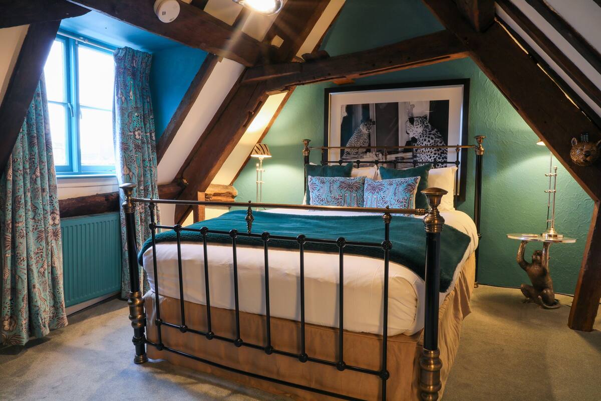A bedroom with a wooden bed, a window with curtains, a framed picture on the wall, and a small table with a lamp.