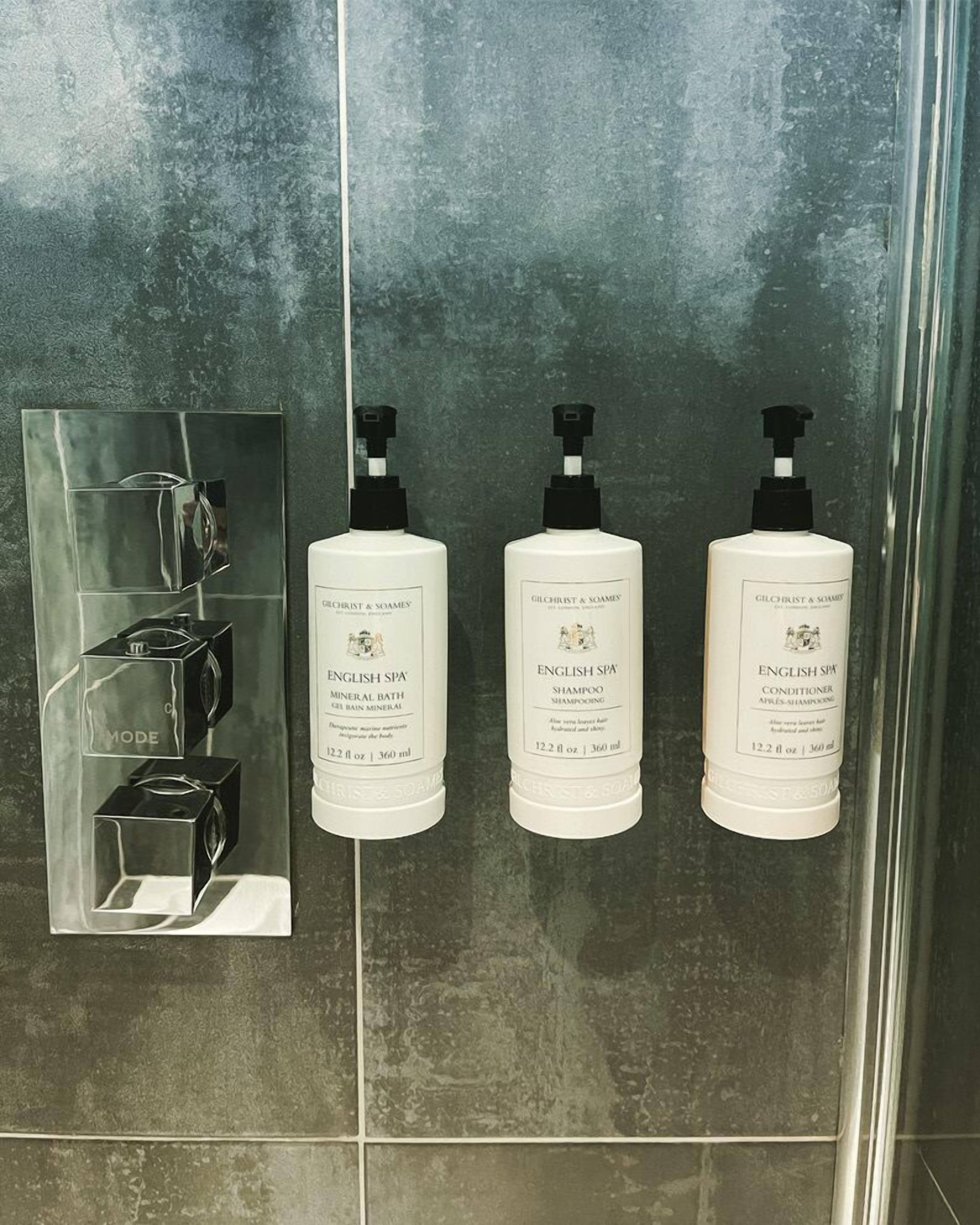 Three bottles of Gilchrist & Soames English Spa Mineral Bath Shampoo and Conditioner on a tiled wall.