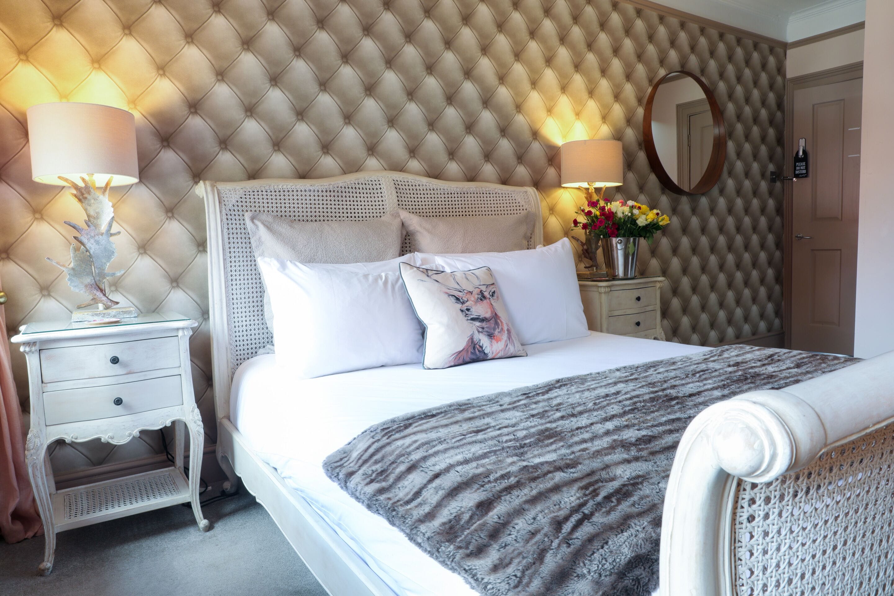 A neatly made bed with a grey blanket, a white headboard, and two bedside tables with lamps and a vase of flowers.