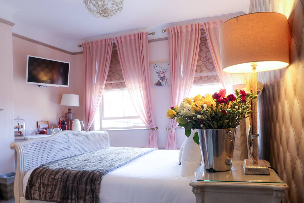 A hotel room with a bed, a bedside table with a lamp, a TV mounted on the wall, and a window with curtains.